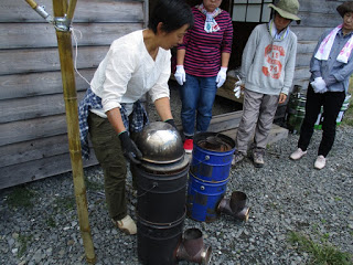 白いシャツの女性がロケットストーブの部品である金属缶を組み立てている様子を周囲の人々が見守っている写真