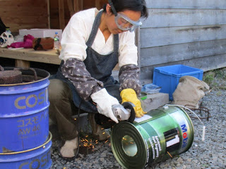 保護メガネと手袋をつけた女性が、電動工具でペール缶を切っている作業中の写真