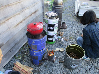 完成したロケットストーブの上に五徳と平鍋が置かれ、隣には赤い鍋をのせた青いペール缶でできたストーブが並べて置かれおり、その前に女性がしゃがんでいる写真