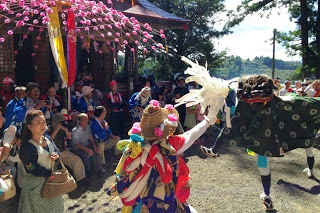 八幡様のお社前に多くの見物客が集まる中、派手な衣装の踊り手が獅子舞と向かい合って踊っている写真