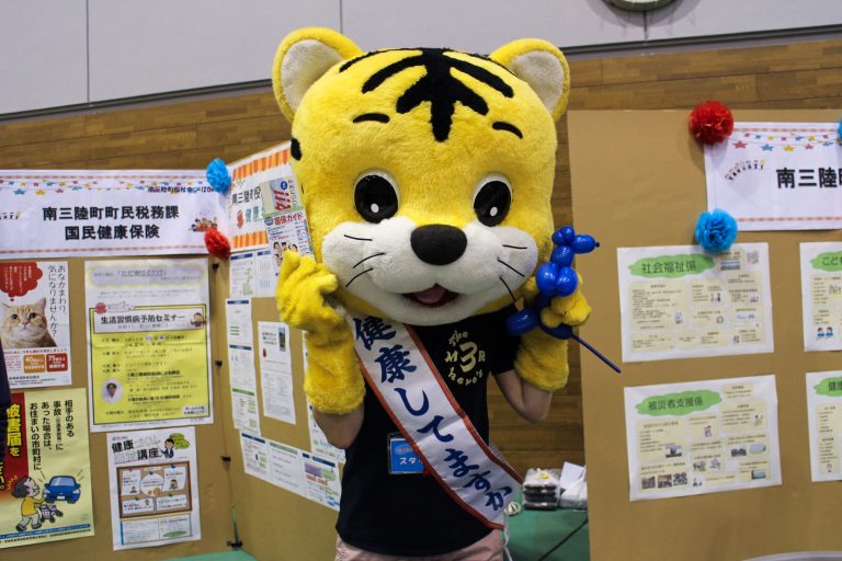 黄色いトラの着ぐるみを着たキャラクターが「健康してますか?」のたすきをしている写真