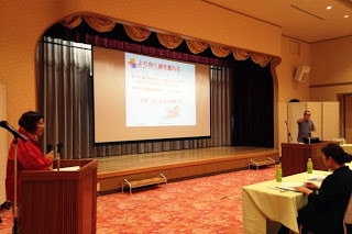 舞台上に資料が映し出されたスクリーンが設置され、左右に司会者と講師が立ち、数人の参加者が机に座っている講演会場の様子の写真