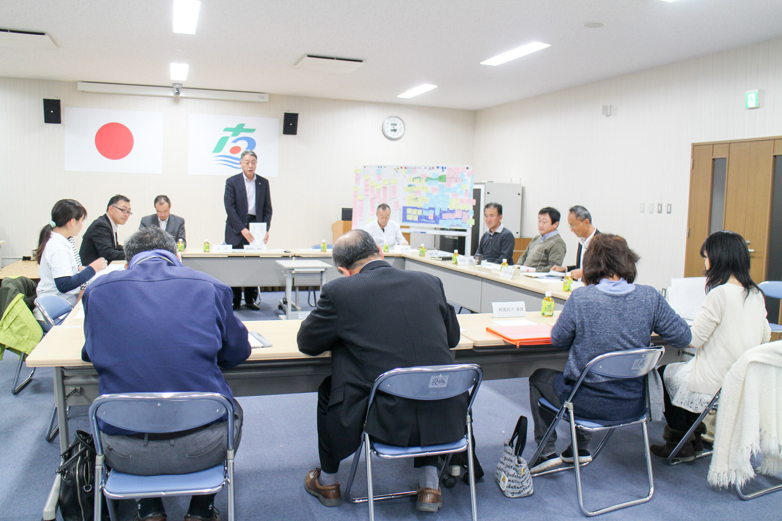 国旗と町旗が掲げられた会議室で、ロの字型に設置されたテーブルの前方に立っているスーツ姿の男性が発言し、他の十数名の参加者が着席し資料を確認しながら真剣に聞いている会議の様子の写真
