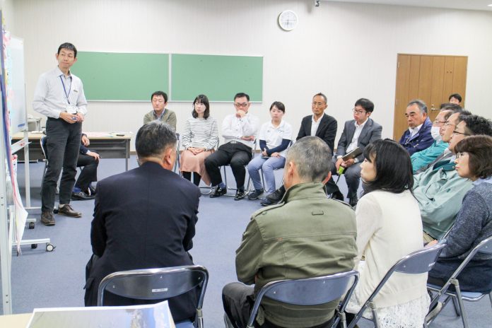 前方に設置されたホワイトボードの前に白いシャツを着た男性が立ち、周囲を囲むように約15人の男女が椅子に座り、手前に座っている男性の話を真剣に聞いている準備委員会の様子の写真