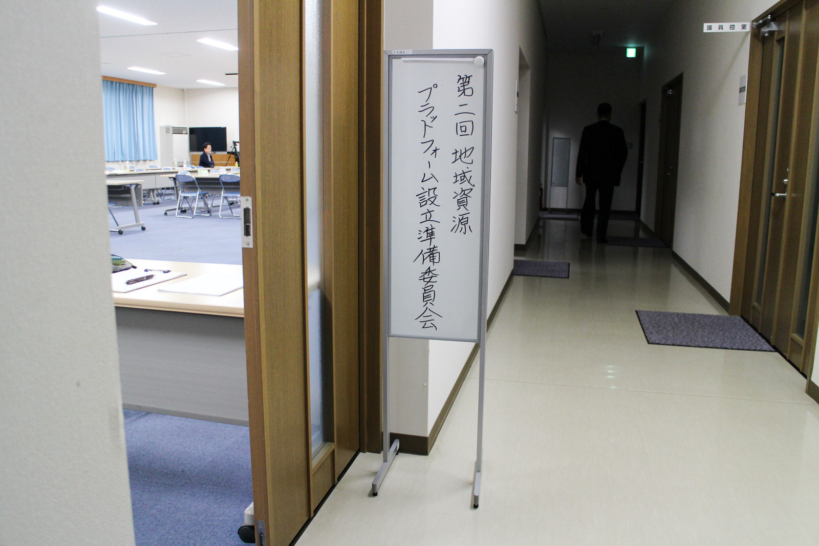 「第二回地域資源プラットフォーム設立準備委員会」と書かれた案内板が廊下に立てられ、その奥にはスーツ姿の男性が歩いていく様子が見える庁舎内の写真