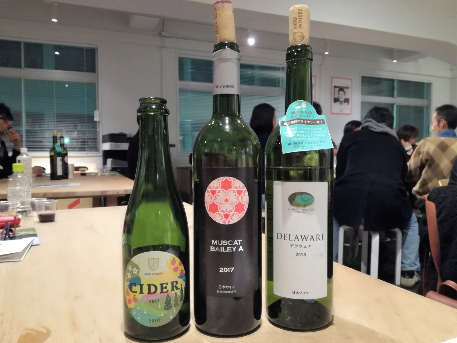 背景には複数の人々が座って談笑している手前のテーブルの上に、ラベルの違う3本のボトルが並んでいる写真