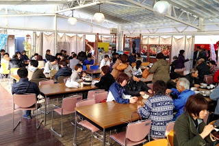 屋根付きの明るい飲食スペースにテーブルが並び、多くの人が座って食事をしている食堂内の写真