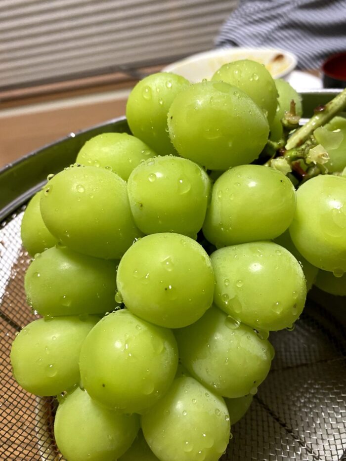 新鮮な緑色のシャインマスカットの房がざるの上に置かれ、果実に水滴がついている写真