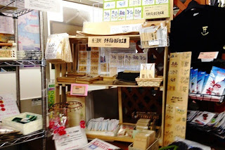 木製の箸・スプーン・しゃもじなどやTシャツ、左側の棚にはパンフレット・地図類、手前に竹細工や木工品が並べられたお土産や地元特産品を販売している場所を写した写真