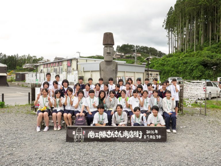 緑豊かな山に囲まれた南三陸さんさん商店街の敷地内で、大きなモアイ像の前にボランティア研修に参加した多くの学生たちが2016年7月23日に集合して撮影した記念写真