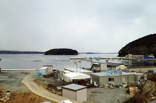 背景には湾と小さな島が見える海に面した場所に、仮設の建物が密集している写真