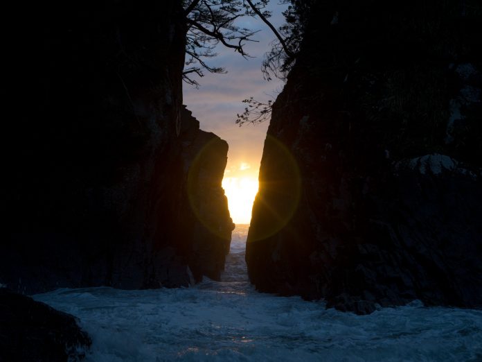左右に分かれた大きな岩の間の狭い隙間から海水が流れこみ、その間からのぼってきた朝日が差し込んでいる神割崎の写真