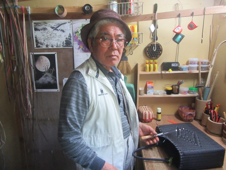 壁にはカレンダーや自然の絵が貼られ、棚には道具や材料が整然と並んでいる室内の工房で、黒い籐のかごを編んでいる帽子を被った佐藤功さんがカメラをみつめている写真