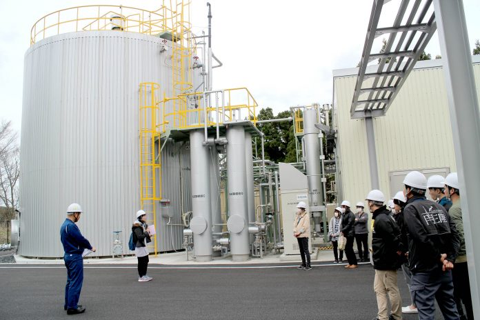 銀色の筒状のタンクや配管が並ぶ工場敷地内で、安全帽をかぶった複数の人が説明を聞いている様子の写真