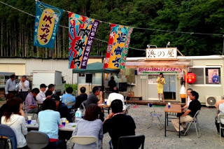屋外に並べられたテーブルと椅子に多くの人が座り、夕方の空の下で女性がステージで歌っている写真