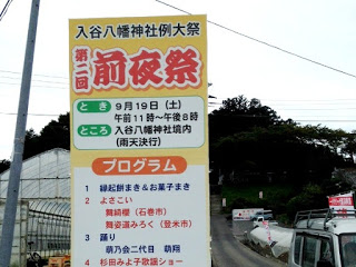 日時や開催場所、催しプログラムなどが大きく書かれている「入谷八幡神社例大祭 第二回前夜祭」の看板が通りに立っている写真