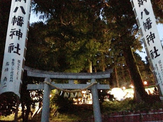 日が暮れかけた山の中で、入谷八幡神社の鳥居の前に白い大きなのぼり旗が立ち、境内に明かりが灯っている写真