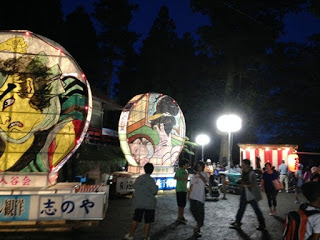 大きなねぶた絵の灯籠が2基明るく照らされて並び、周囲では家族連れや子どもたちが歩き回り、奥には赤白幕の舞台が見える夜の神社境内の写真