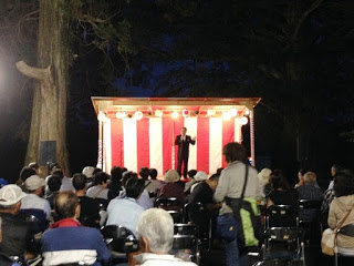 赤白幕で飾られた小さな舞台の上でスーツ姿の男性が話しており、その前に多くの観客が椅子に座って真剣に耳を傾けている写真