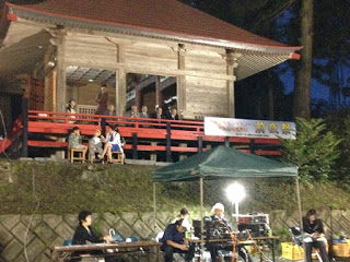 神社の拝殿に数人が座り見物している様子と、その手前でスタッフらしき人たちが音響や照明機材を操作している様子が写っている写真