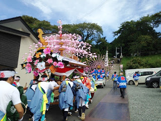 男性たちがピンク色の花飾りを付けた大きな山車を力を合わせて坂道の上へ引っ張っている祭りの様子の写真