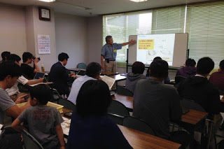 教室内でスライドを使いながら講師の男性が化石に関する説明をしており、机に座った参加者たちが真剣に話を聞いている様子の写真