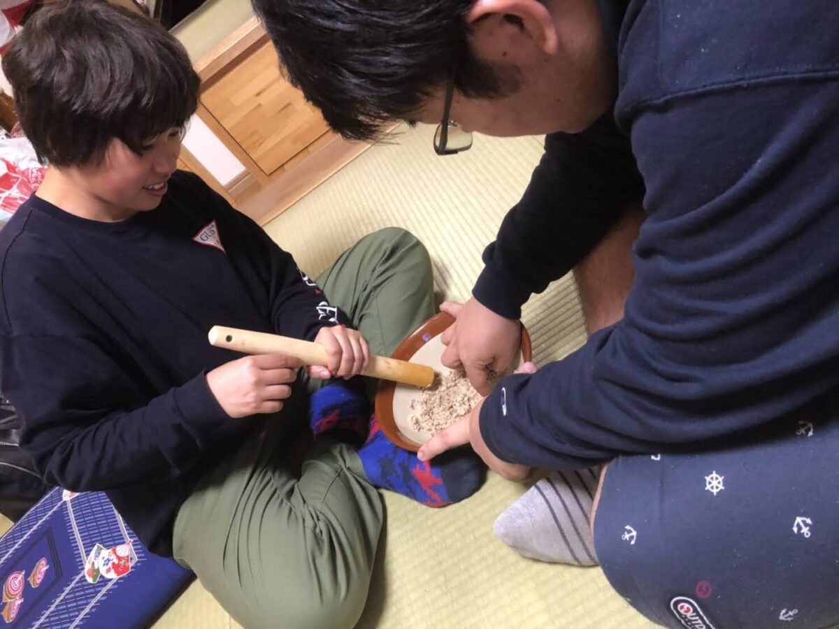 畳の上に向かい合って座る二人の人物が、一人がすりこぎを持ち、もう一人がすり鉢を押さえながら協力してくるみをすり潰している作業風景の写真