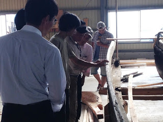 屋内の作業場で複数の男性が古い木製の船の一部を囲んで修復の説明を受けながら観察している様子の写真