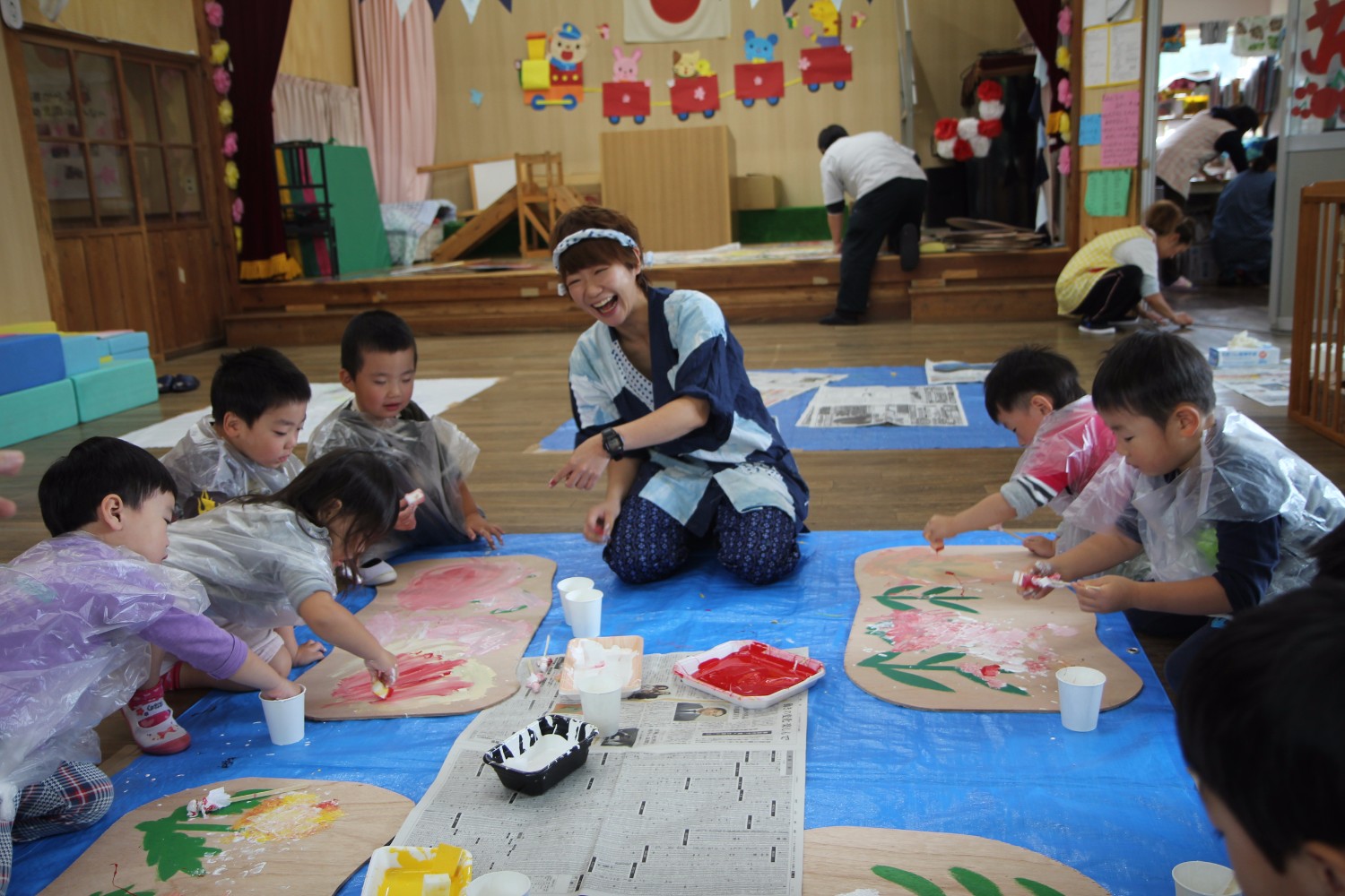 幼児園の室内で子どもたちがビニール製のエプロンを着て、床に置いた大きな紙に絵の具で自由に絵を描いており、中央でYORIKOさんが笑顔で見守っている創作活動の写真