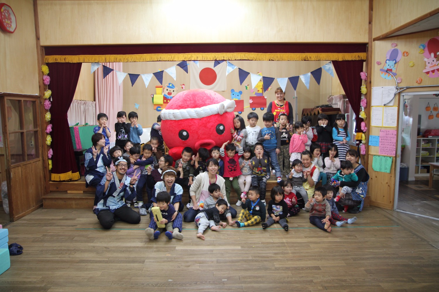 舞台の前に集まった大勢の園児と保育士、オクトパス君、藤田岳さんとYORIKOさんが一緒に集まって笑顔で記念撮影をしている集合写真