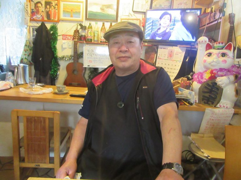 木製のカウンターと、大きな招き猫などの飾りに囲まれた店内で、帽子をかぶった渡辺清吾さんが椅子に座っている写真