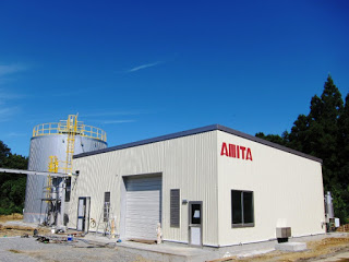 青空の下、AMITAと書かれた白い建物と大型タンクが立ち並び、周囲は自然に囲まれているバイオガス施設の外観の写真