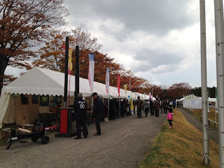 紅葉の木々が並ぶ屋外の通りに白いテントが連なり、多くの人が歩いたり商品を見たりしている様子が写っている写真