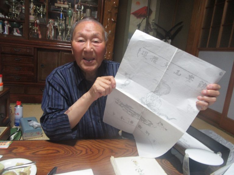 掛け軸や沢山のトロフィーが飾られた和ダンスが置かれた畳の和室で、小野實さんがイラストが描かれた大きな紙を広げて見せている写真