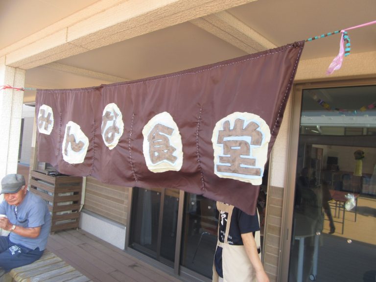 建物の軒先に「みんな食堂」と書かれた布看板が吊るされ、左側のベンチには高齢の男性が腰掛けている食堂入口の写真