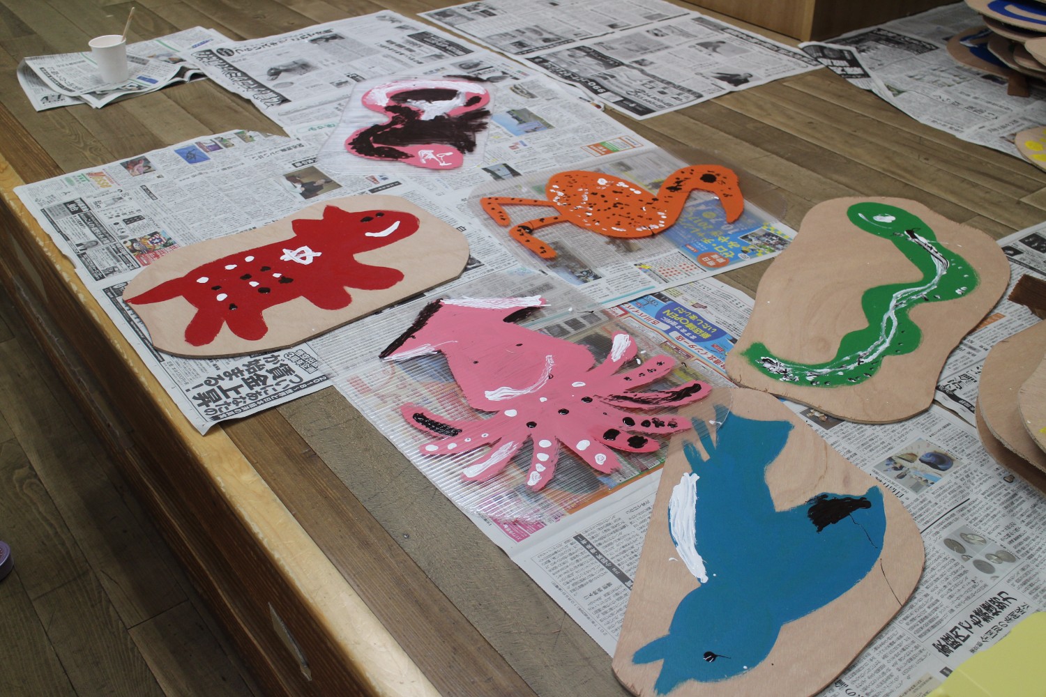 新聞紙の上に並べられた木の板に、子どもたちが描いたイカやトリ、ヘビなどのカラフルな動物や生き物の絵が乾かされている作品展示の写真