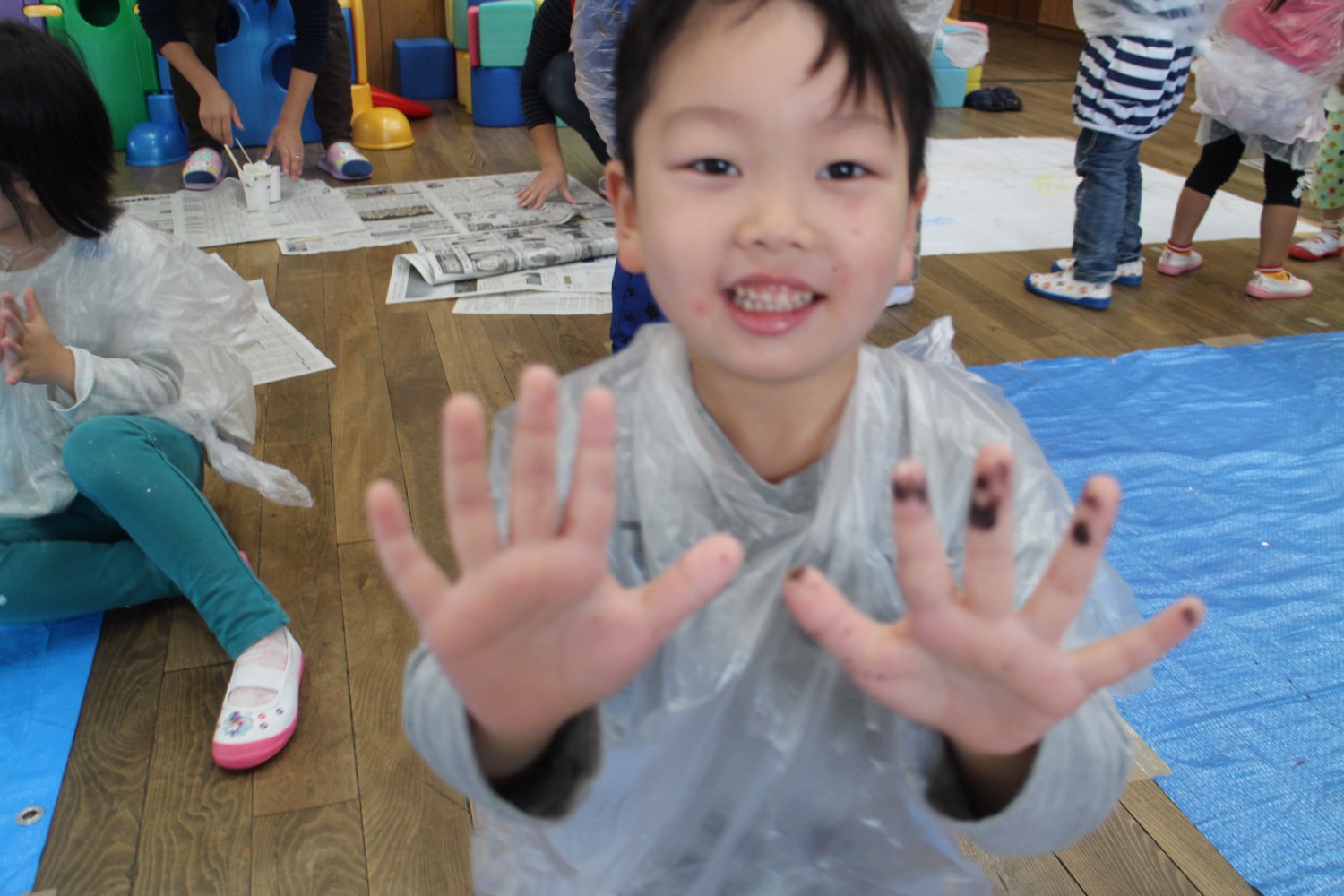 絵の具のついた両手を広げて笑顔でカメラに近づく男の子が写っており、後ろには他の子どもたちが創作活動を楽しんでいる室内の様子の写真