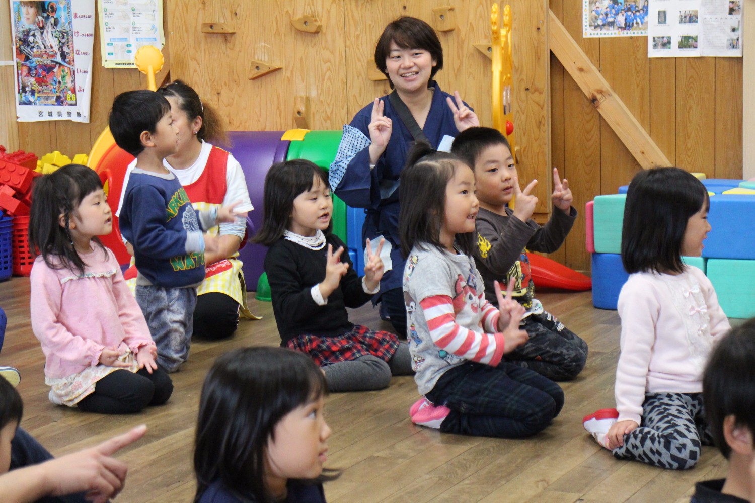 子どもたちが保育士が一緒に座り、両手で指を立ててリズム遊びを楽しんでいる様子の写真