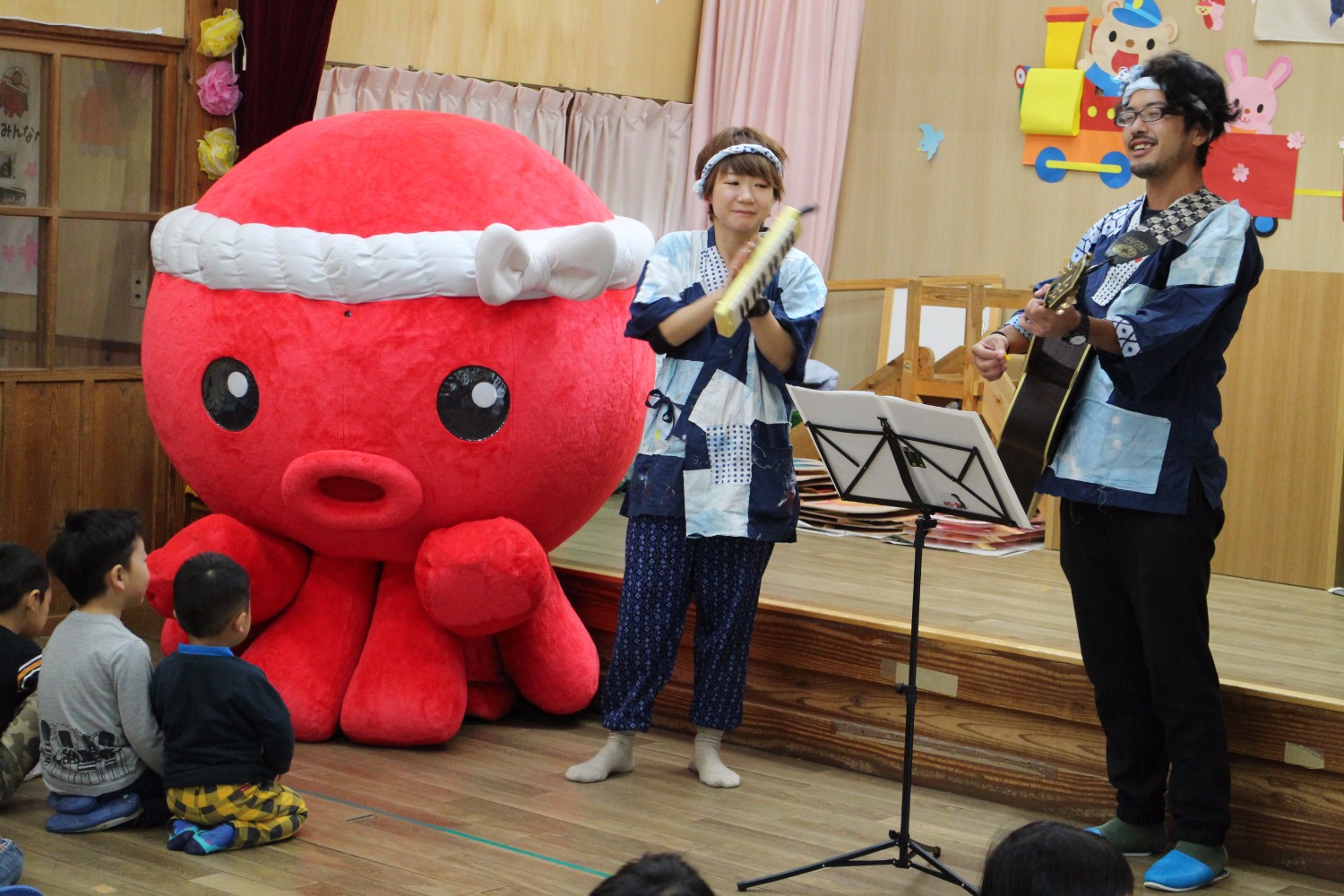 幼児園のステージの前で、大きな赤いタコの着ぐるみキャラクターオクトパス君と並んで、法被姿の藤田岳さんとYORIKOさんがギターと鍵盤ハーモニカの演奏で子どもたちに楽しい出し物を披露している写真
