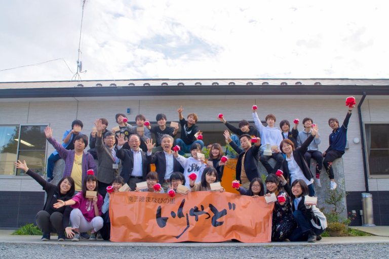 白い外壁の住宅の前で、老若男女が大きな橙色の横断幕を持ちながら、赤い物を手に全員で笑顔を見せている集合写真