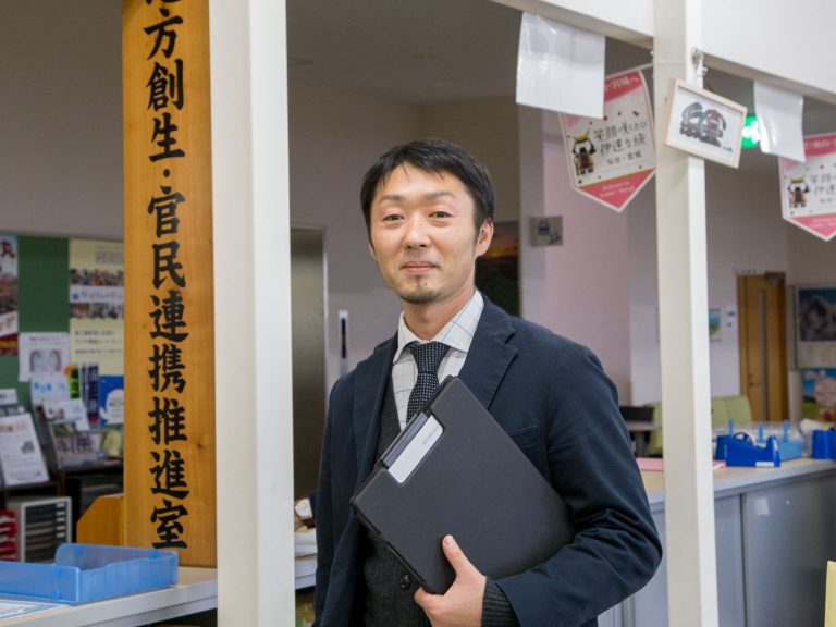 スーツを着た男性が「官民連携推進室」と書かれた看板の横でクリップファイルを持って笑顔で立っている阿部大輔さんをアップで撮影した写真