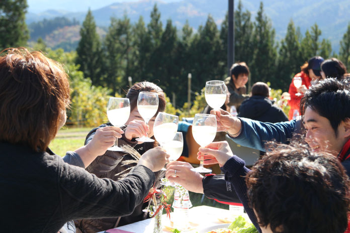 山や森に囲まれた自然の中、屋外のテーブルを囲んだグループの参加者達が、ワインが入ったワイングラスを持ち上げて、みんなで乾杯している様子の写真