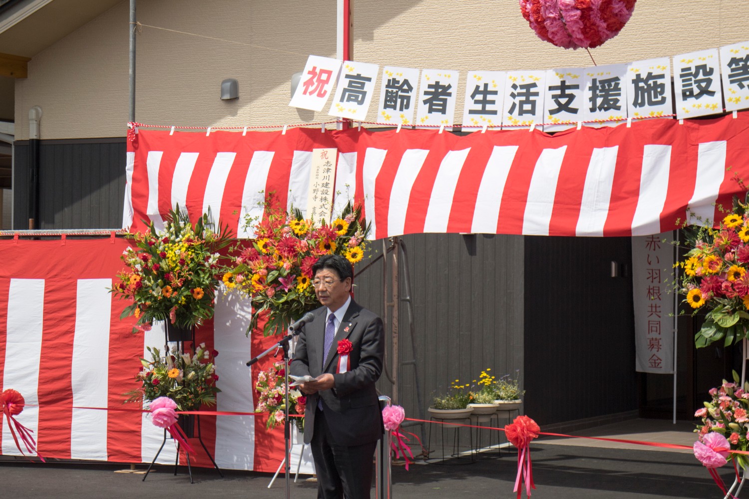 背景に横断幕や紅白幕、祝い花が飾られた建物前で、スーツ姿の佐藤町長がマイクの前に立ち挨拶を行っている写真