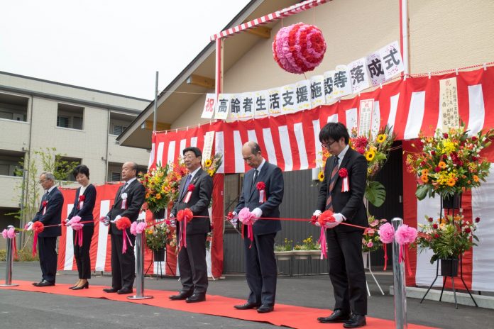 「高齢者生活支援施設等落成式」と書かれた横断幕やくす玉が設置され、お祝い花が飾られ紅白幕が貼られた建物入り口前に、敷かれたレッドカーペット上に立つ地元の関係者が紅白のテープを手に並んでいるテープカットの様子の写真
