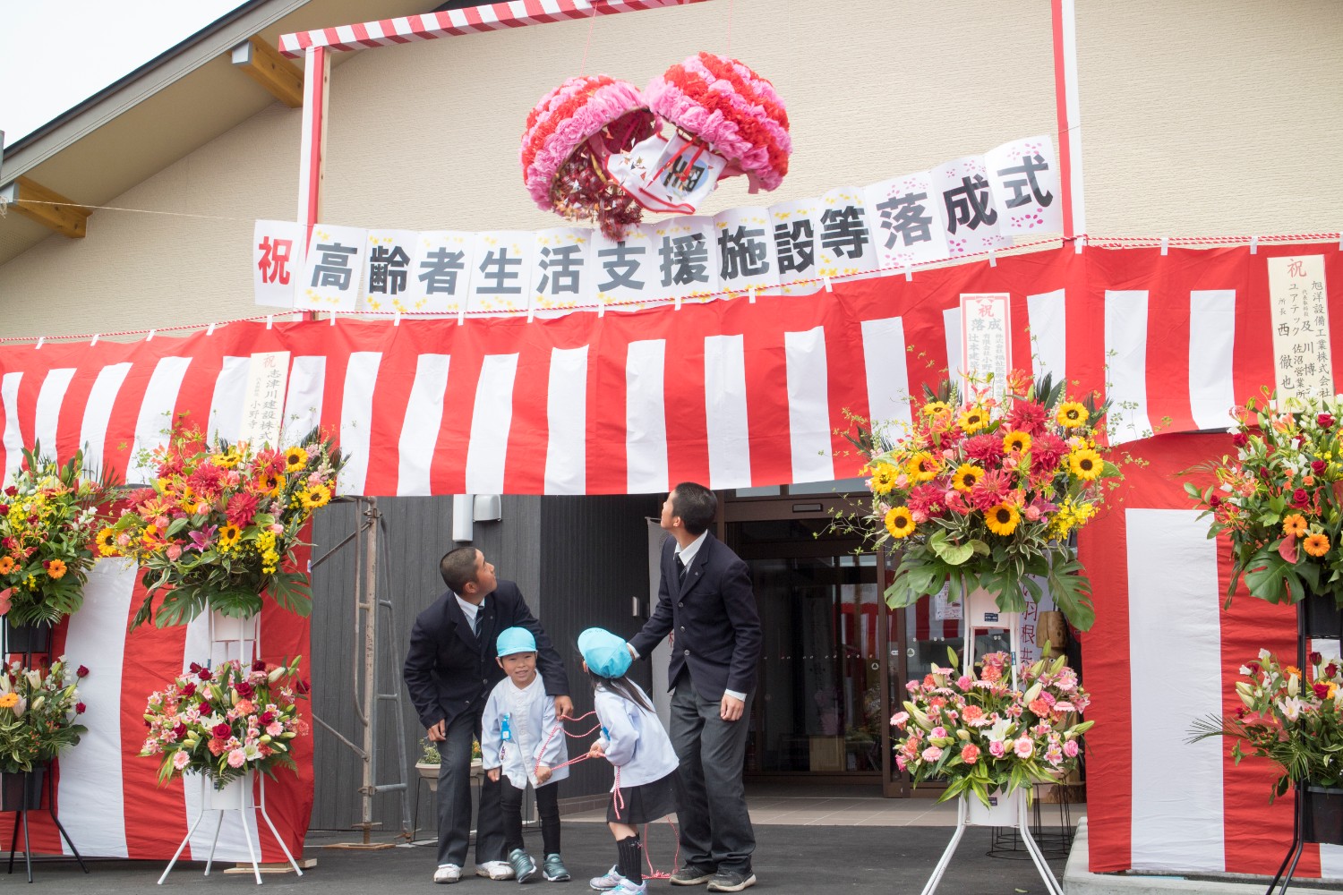 横断幕や紅白幕が貼られ、祝い花が飾られた正面玄関前で、紐を引っ張り上を見上げている2人の園児と2人の男子生徒の頭上で、ピンクと赤色のくす玉が割られている瞬間の様子の写真