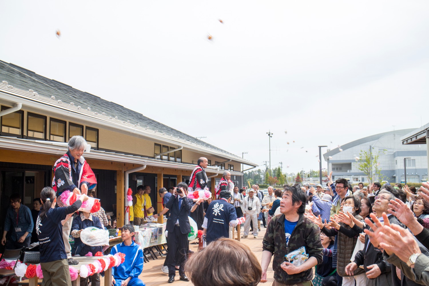 屋外広場で壇上の3人が小さなお餅を投げ、集まった来場者たちが上を見上げ楽しそうに受け取ろうとしている写真