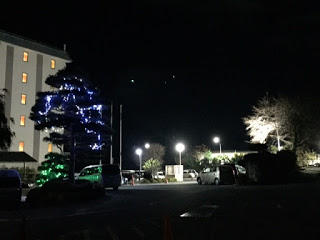 夜の駐車場に複数の車が停められており、左側の建物の窓からは室内の明かりが漏れ、近くの木には青と緑のライトが飾られ、奥の木には白いライトが灯り、街灯が駐車場全体を照らしている様子の写真