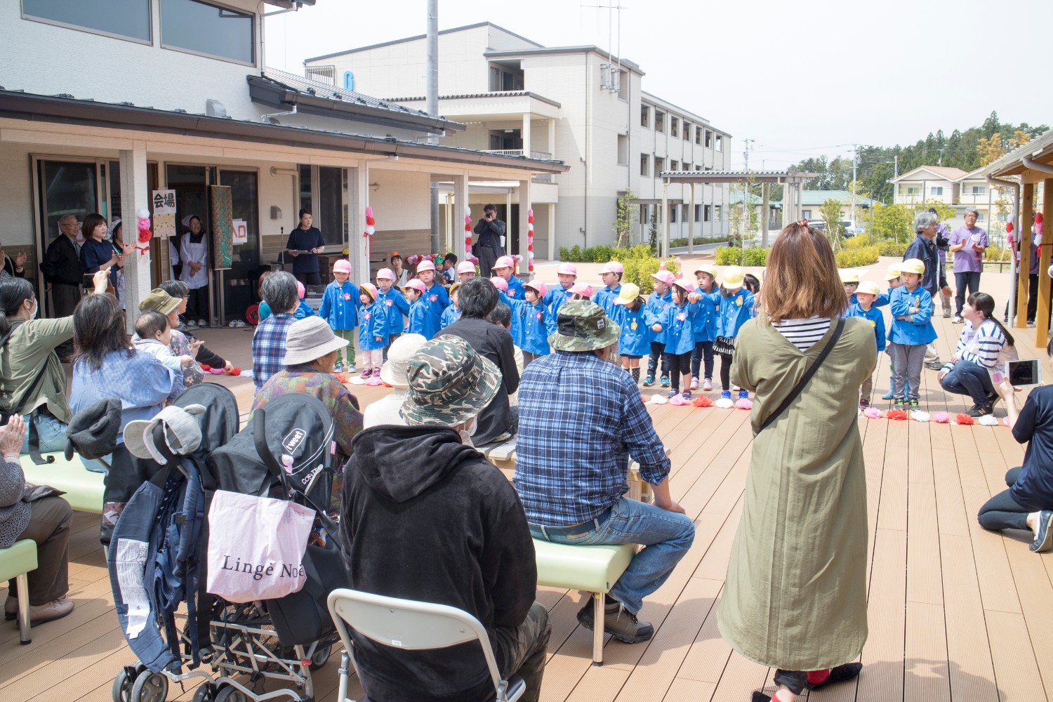 屋外広場の長椅子やパイプ椅子に座った人達の前で、ピンク色や黄色の帽子に青色の制服を着た園児たちが2列に並んで踊りを披露している写真