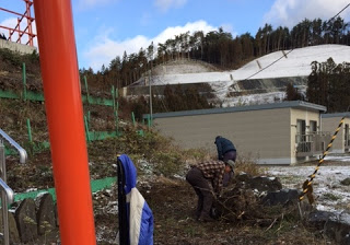 朱色の鳥居が立つ雪の積もった屋外で、斜面と建物を背景に、厚着をした二人の人物が地面の草や枝を集めて神社の掃除をしている様子の写真