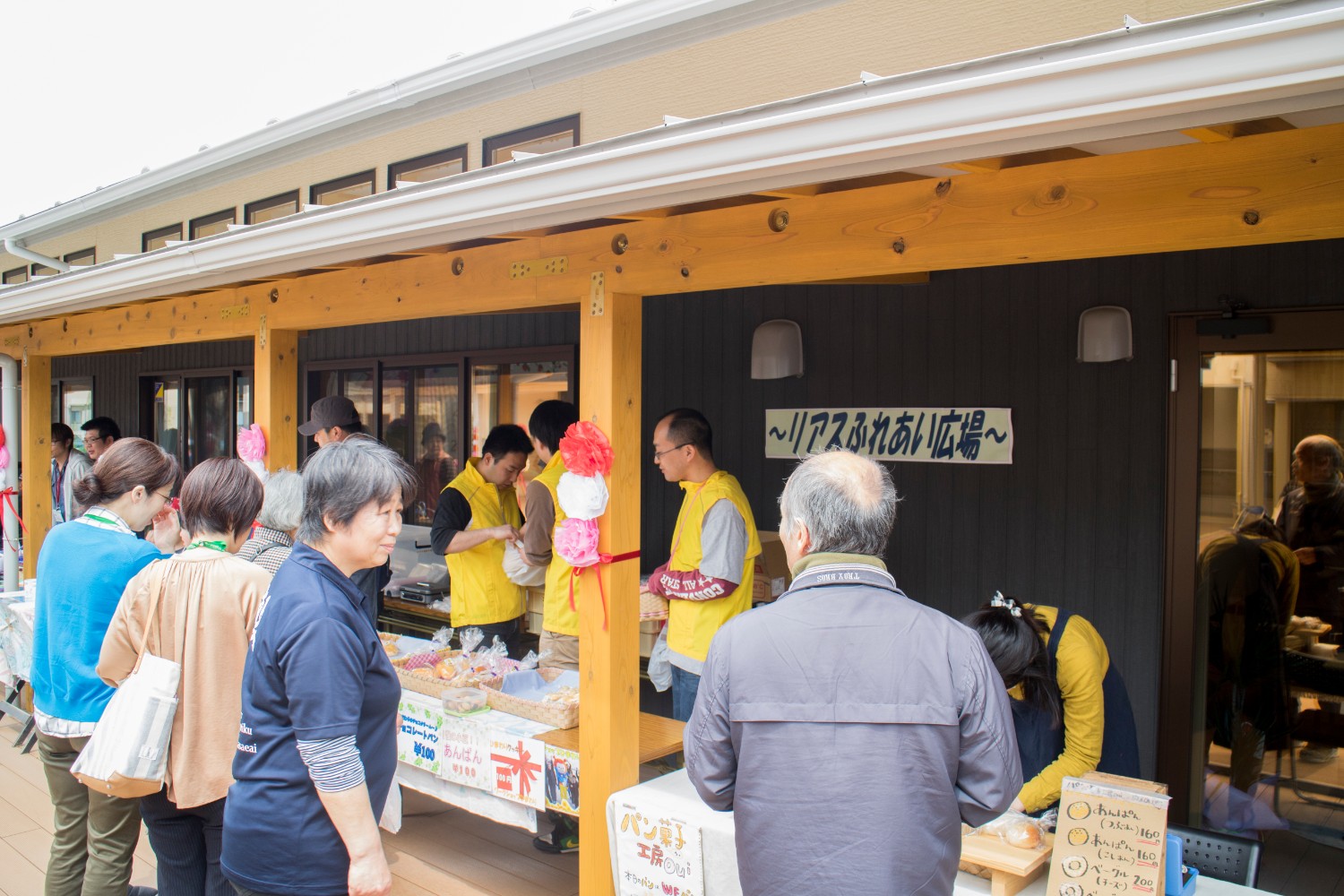 建物の壁に「リアスふれあい広場」と書かれた屋外の軒下で、黄色いベストを着たスタッフが屋台でパンを販売している会場で来場者が買い物を楽しんでいる様子の写真